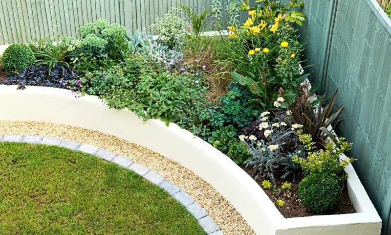 Curved garden bed with diverse colorful plants, surrounded by a gravel path and bordered by a green fence