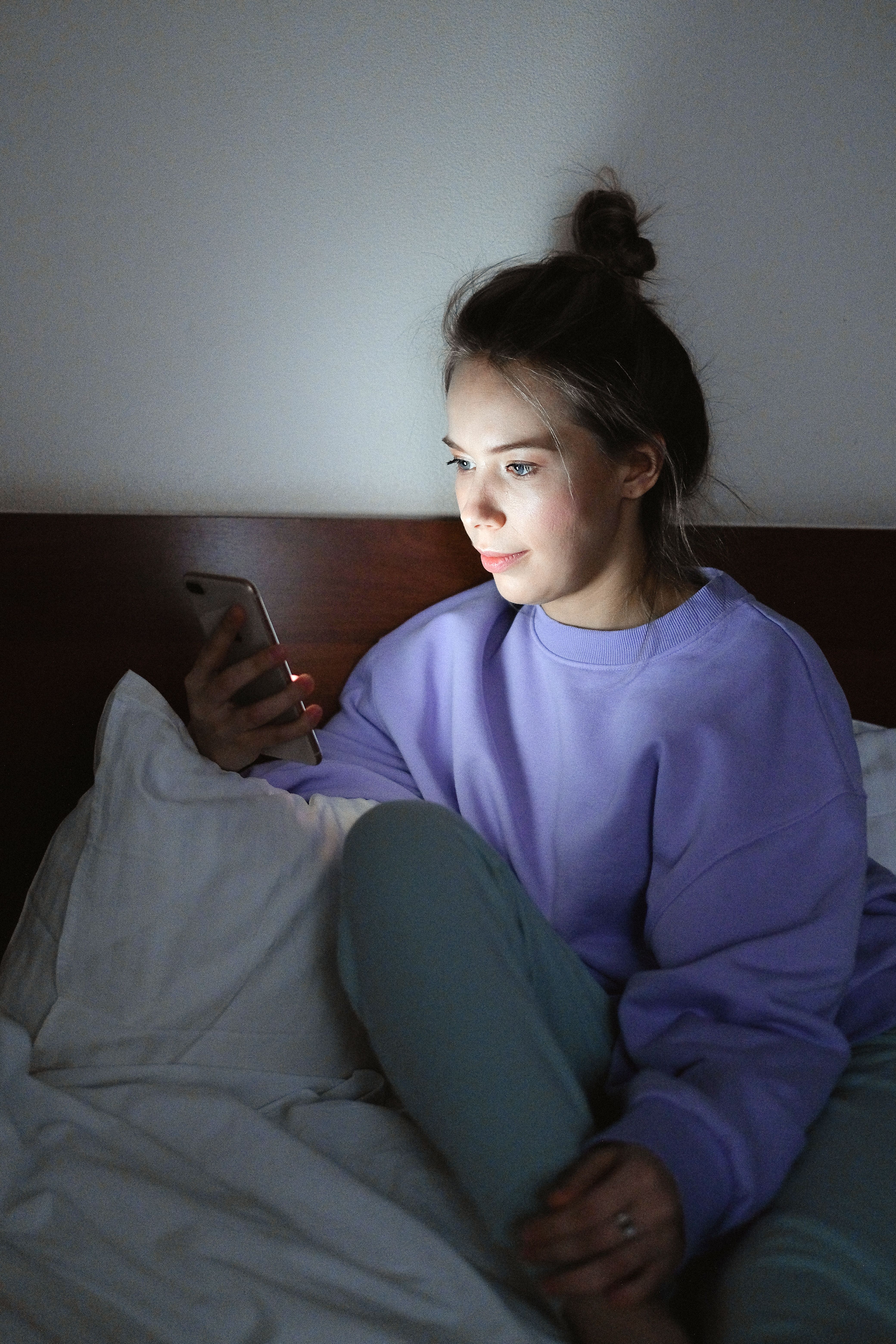 Young woman lying alone in a dimly lit bedroom scrolling her smartphone at night
