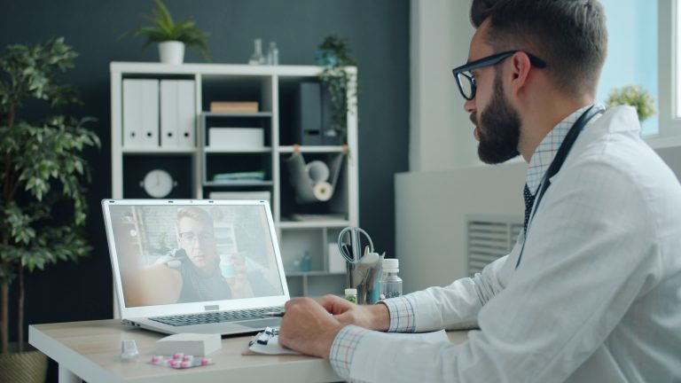 Doctor in white coat video calling a patient on a laptop in a modern office setting