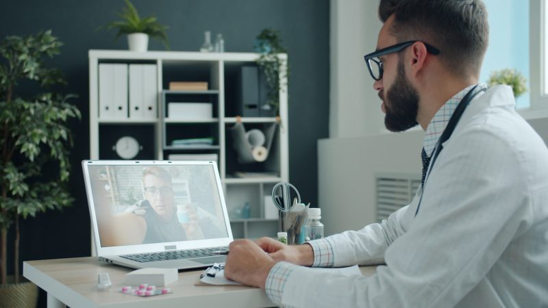 Doctor in white coat video calling a patient on a laptop in a modern office setting